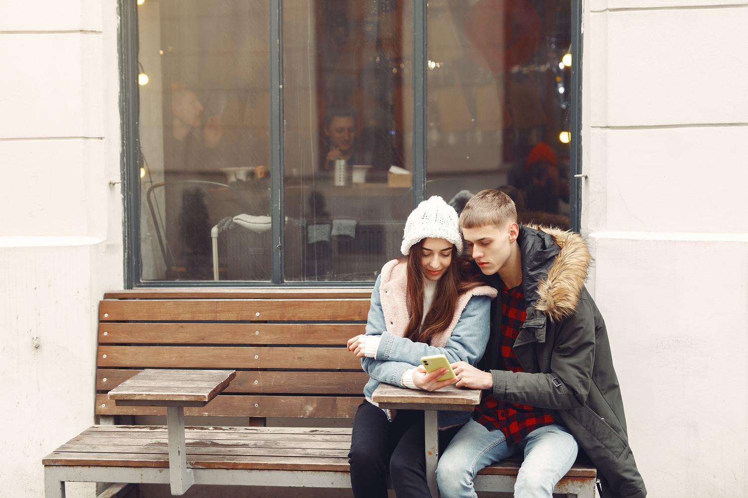 Una pareja joven sentada en un banco revisando juntos un teléfono móvil, representando una cita exitosa.