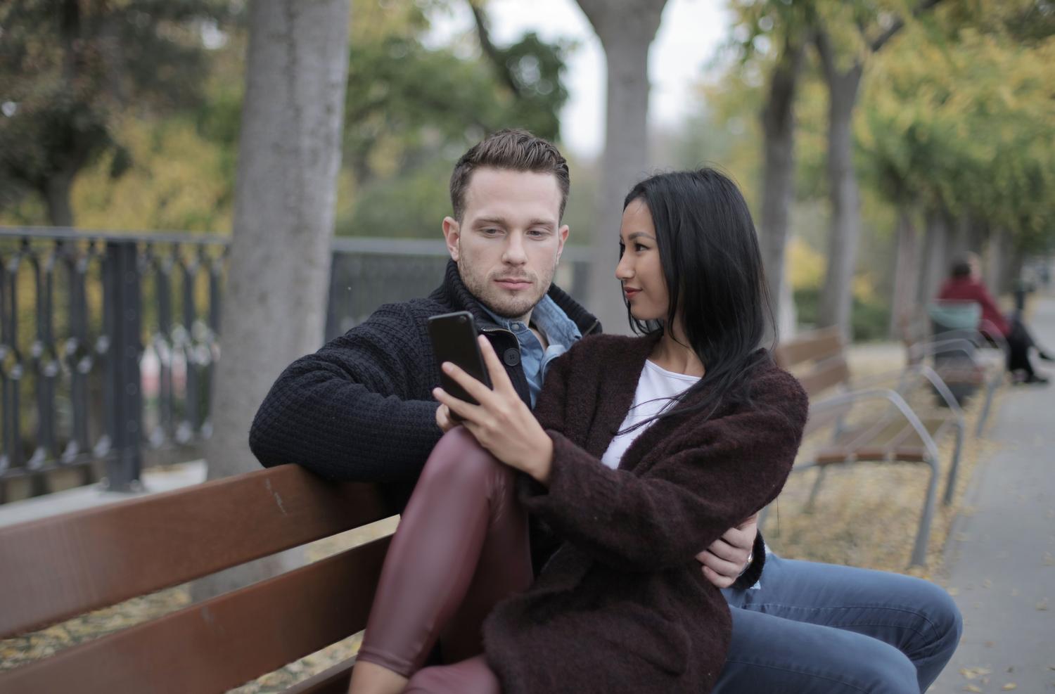 Pareja multirracial sonriendo mientras usan un smartphone en un parque, ilustrando la conexión a través de la tecnología.