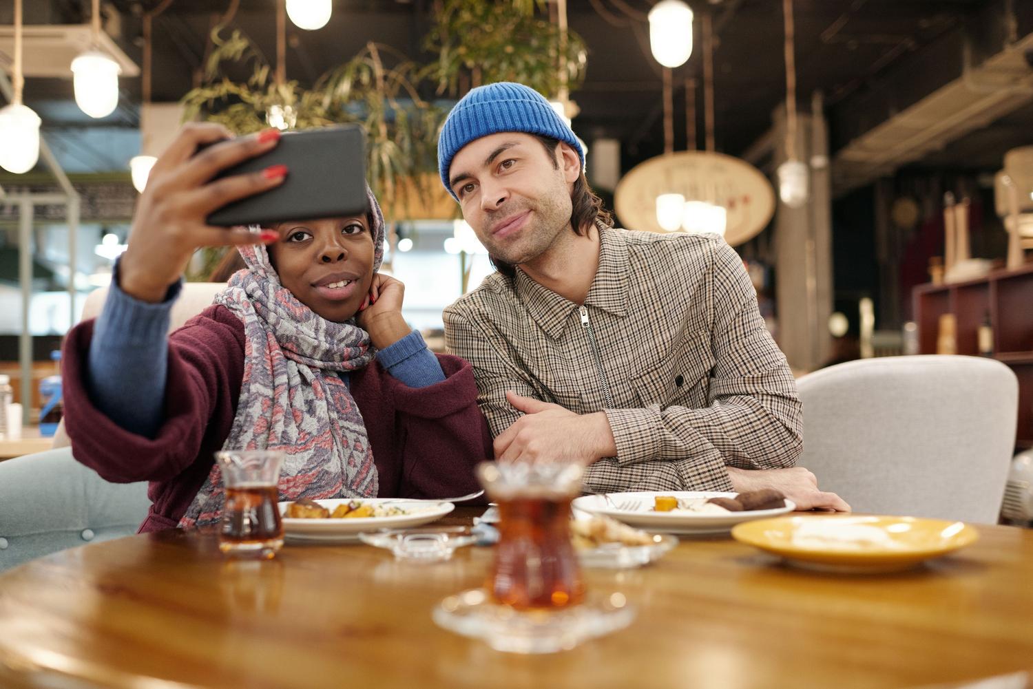 Pareja feliz tomándose una selfie en un café moderno, simbolizando el éxito de conocer a alguien mediante apps de citas.