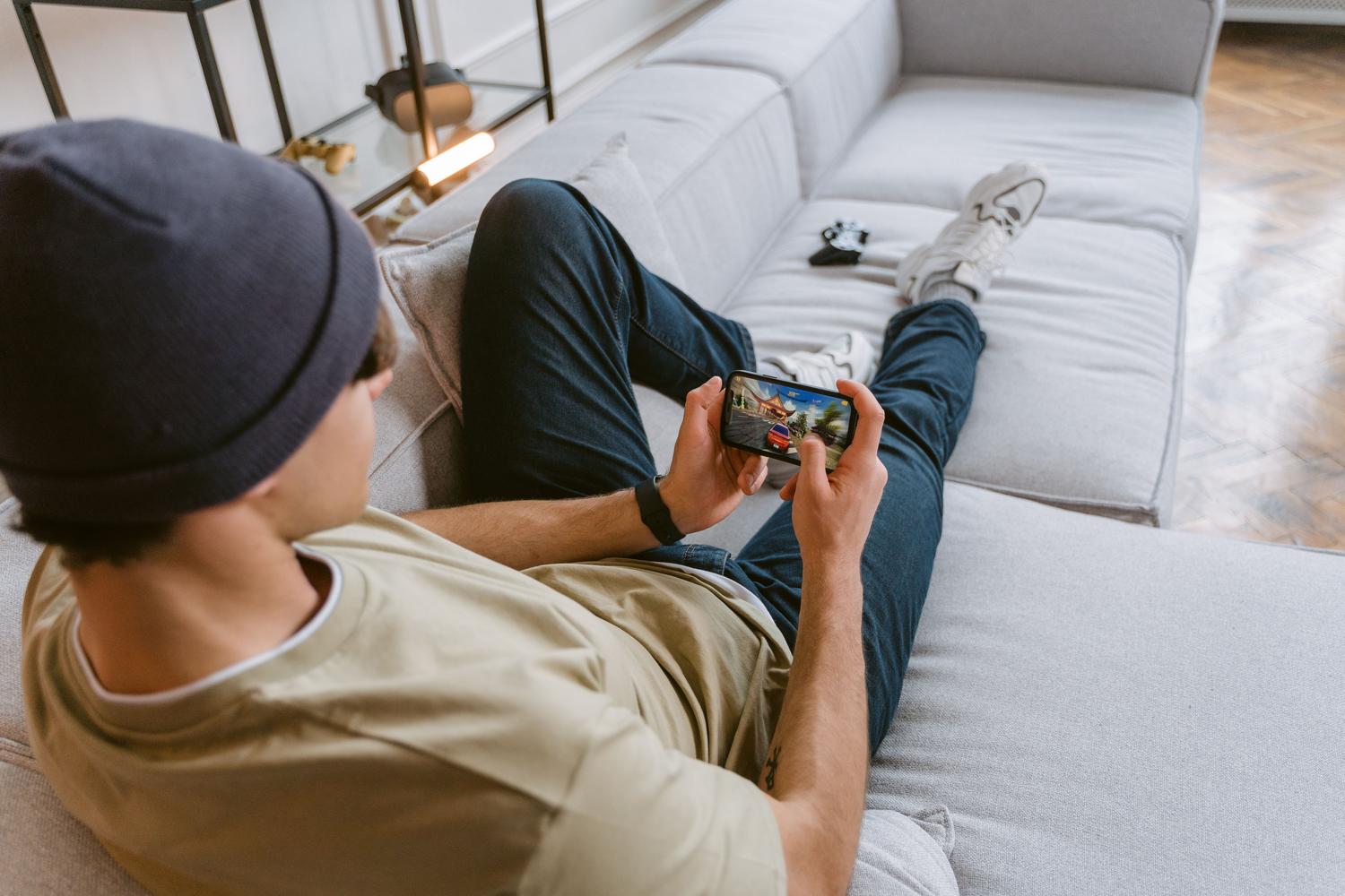 Joven relajado en el sofá jugando a un videojuego en su smartphone, ilustrando la experiencia de juego de Heartopia