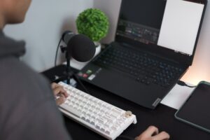 Persona sentada en un escritorio moderno con un portátil, un micrófono USB y un teclado, en un setup de grabación casera para streaming o podcasting