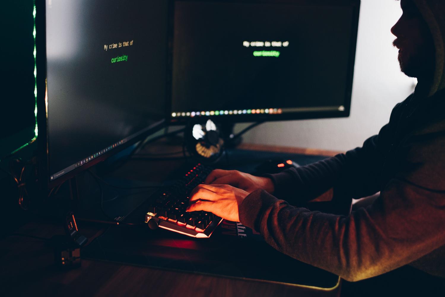 Silueta de un hacker escribiendo en un ordenador en una habitación oscura con pantallas que muestran código verde, representando la amenaza de los virus informáticos.