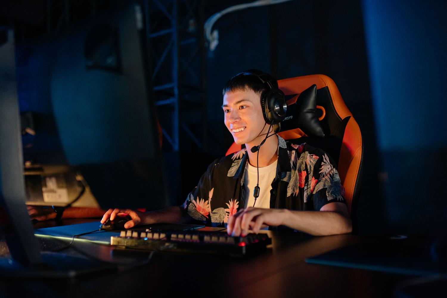 Hombre joven sonriendo mientras juega en línea con auriculares y teclado retroiluminado.