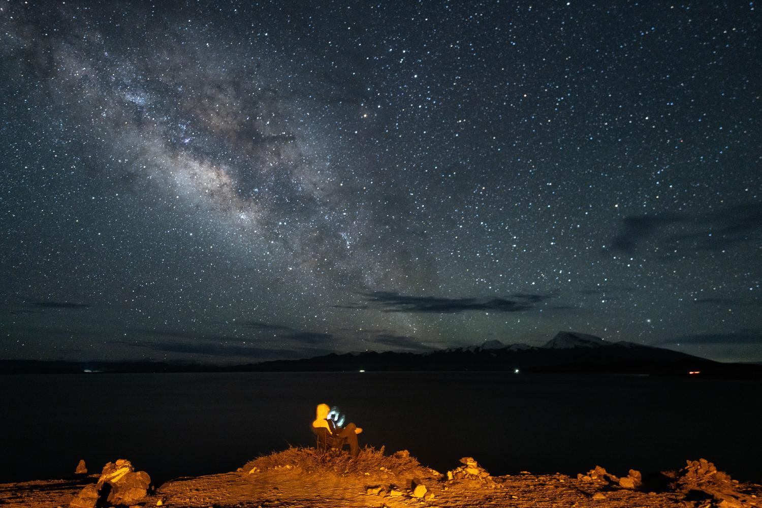 Persona observando las estrellas bajo la Vía Láctea en una noche despejada junto a un lago
