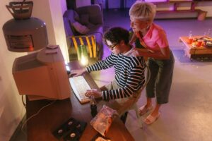 Două persoane folosesc un computer vechi cu monitor CRT, evocând nostalgia pentru jocurile pe PC din anii '90.