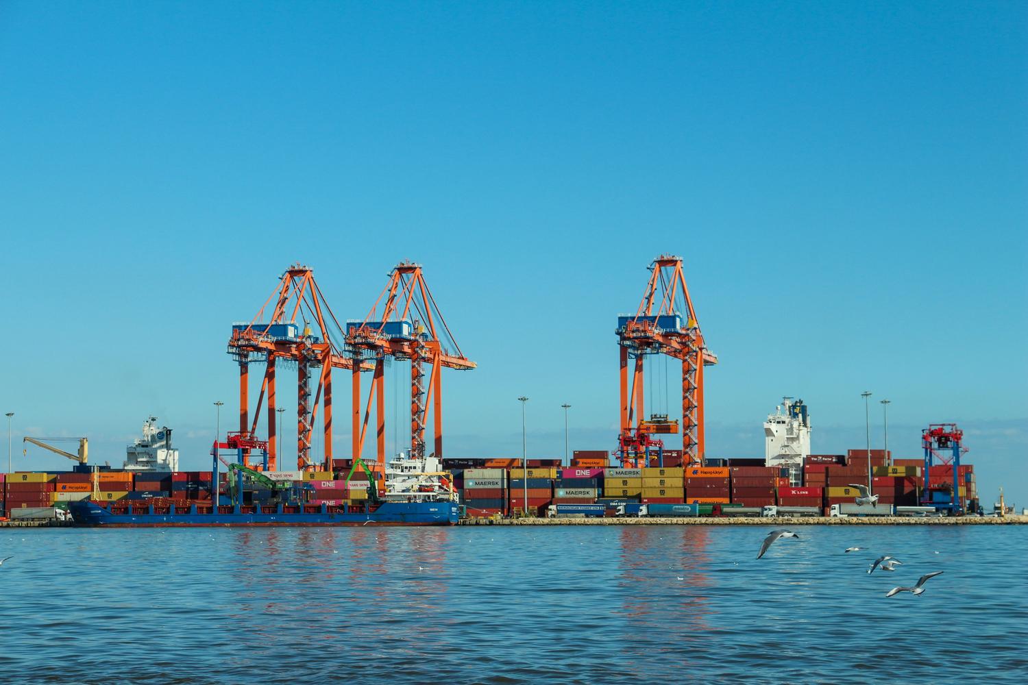 Industrial shipping port with large orange cranes and colorful containers, representing Port Gellhorn. Industrial shipping port with large orange cranes and colorful containers, representing Port Gellhorn.