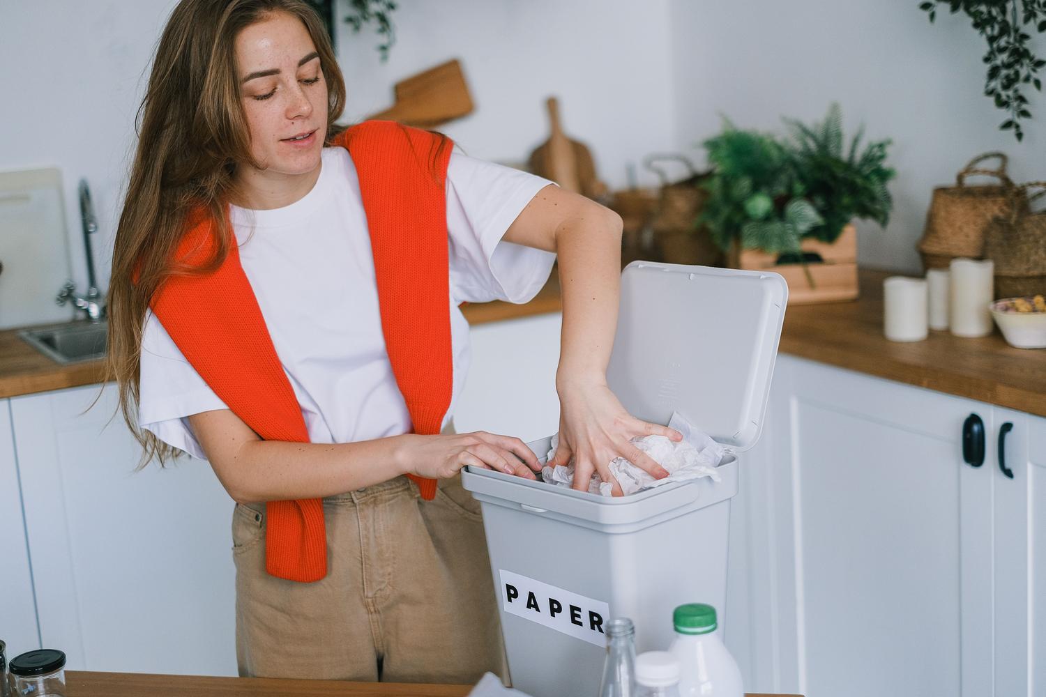 Una mujer joven separando residuos y colocando papel en un cubo de plástico en una cocina iluminada.