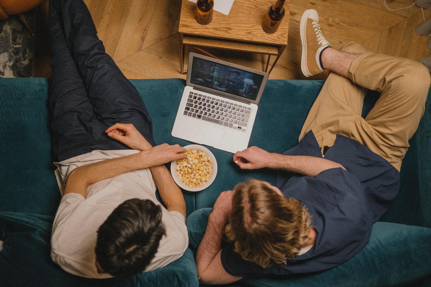 Dos amigos relajados en un sofá viendo una serie en un ordenador portátil con palomitas, simulando un maratón de anime en Netflix.