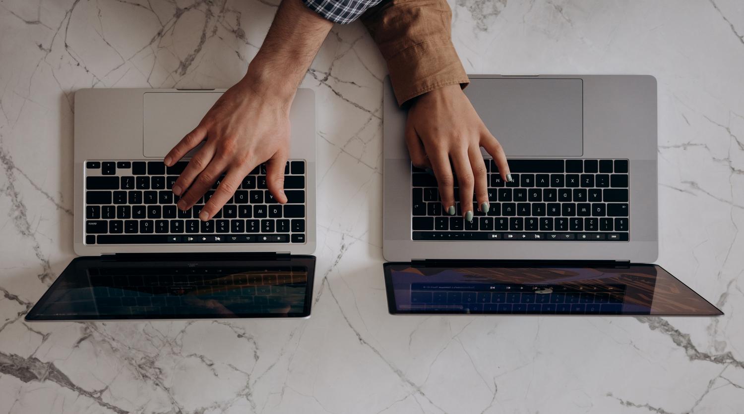 Vista superior de dos personas trabajando con portátiles simultáneamente, representando la sincronización y colaboración entre múltiples dispositivos.