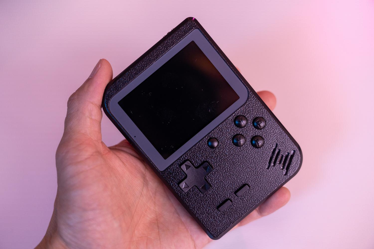 Consola portátil retro de estilo Game Boy sostenida por una mano sobre un fondo rosa vibrante.