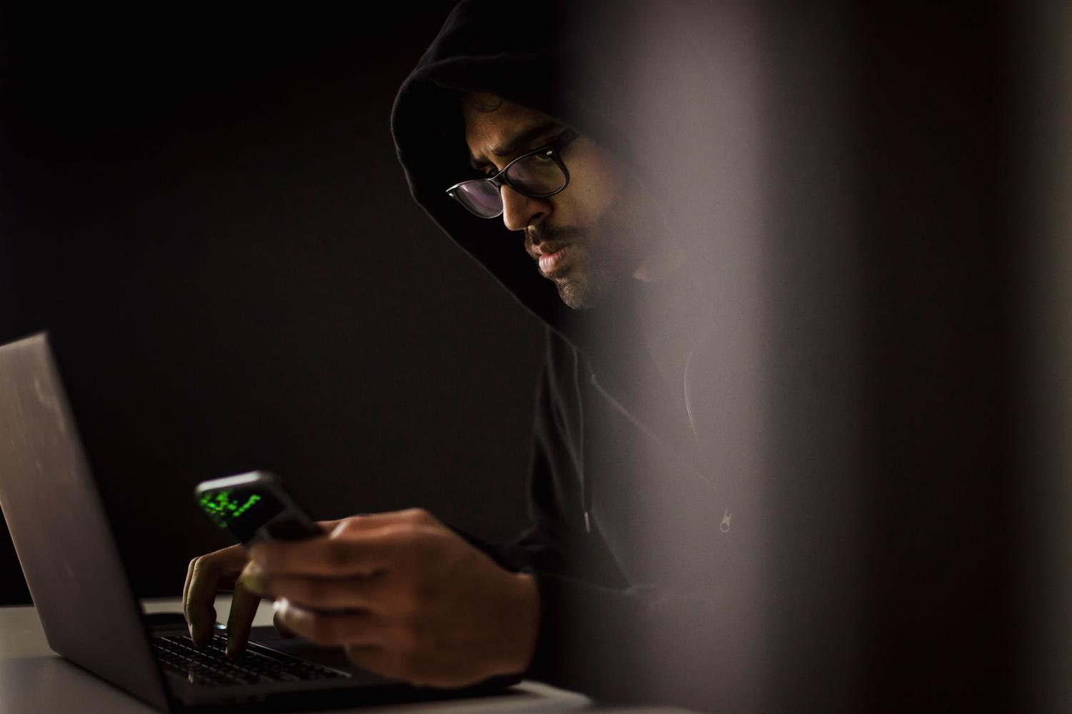 Ciberdelincuente con capucha utilizando un smartphone y un portátil en un entorno oscuro.