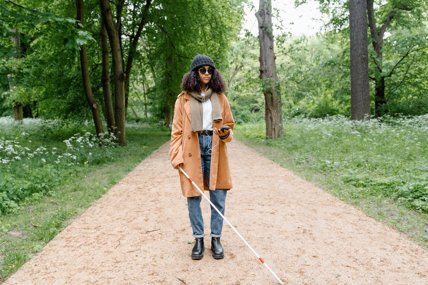 Mujer con discapacidad visual utilizando un smartphone y auriculares mientras camina con su bastón blanco por un parque, ilustrando la autonomía y el apoyo tecnológico en la movilidad.