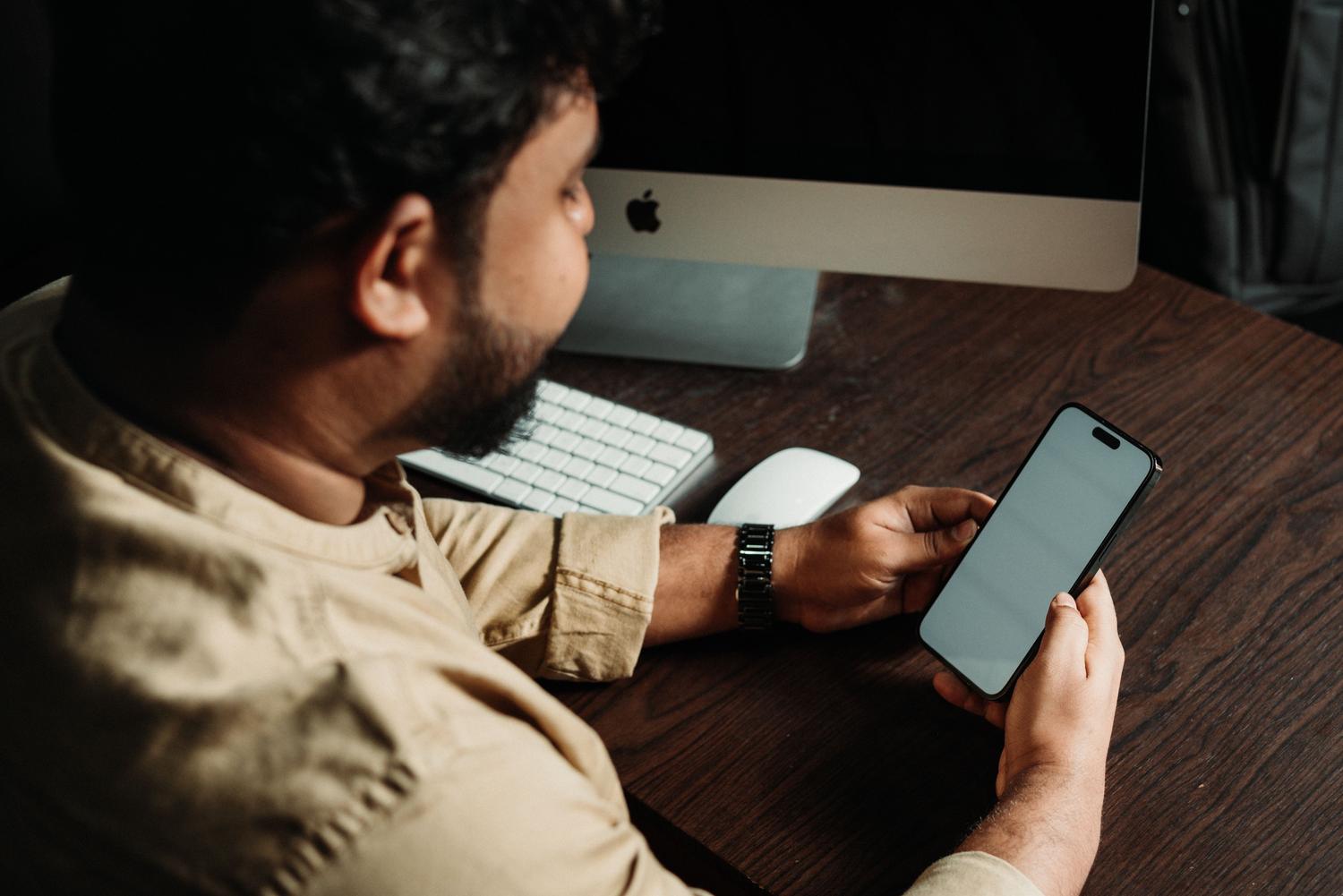 Trabajador de oficina utilizando su iPhone junto a un ordenador iMac, mostrando un entorno de trabajo tecnológico.