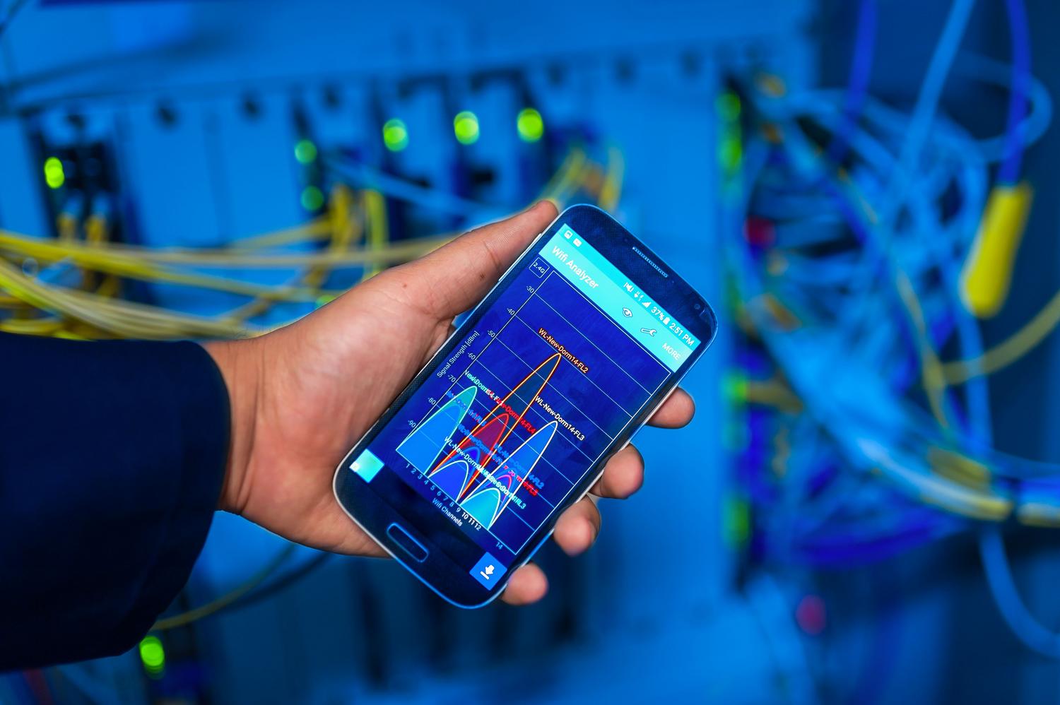 Técnico utilizando una aplicación de análisis de señal WiFi en un smartphone dentro de un centro de datos con racks de servidores, representando la medición técnica de la velocidad y calidad de red.