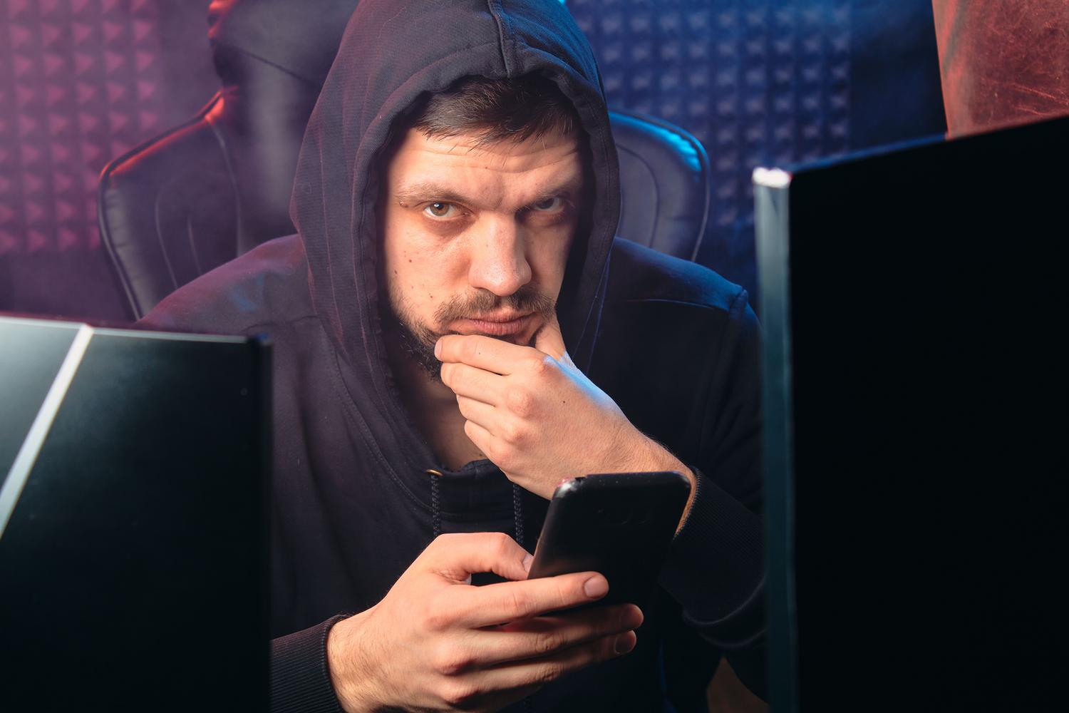 Hacker con capucha utilizando un smartphone en un entorno digital oscuro, representando al estafador.