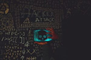Hacker con capucha utilizando una tablet que muestra una calavera, rodeado de símbolos de ciberataque en una pizarra.