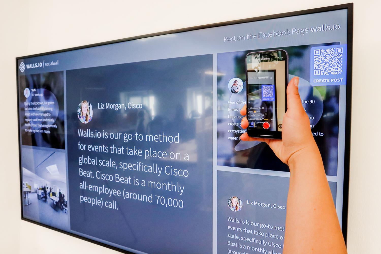 Mano sosteniendo un smartphone para escanear un código QR en una pantalla digital grande, ideal para ilustrar el uso de QRs en publicidad y eventos.