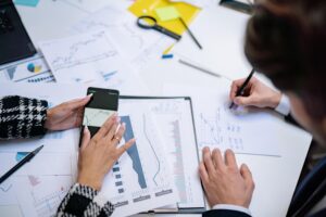 Dos profesionales analizando gráficos financieros y datos con un smartphone y documentos sobre una mesa.