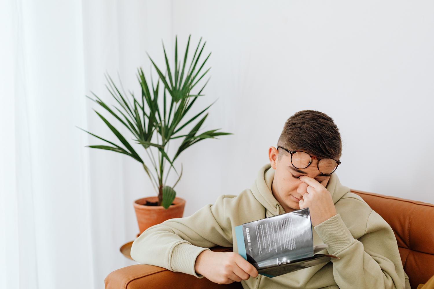 Hombre adulto frotándose los ojos mientras lee un libro, mostrando la fatiga y el cansancio extremo que puede persistir en la edad adulta