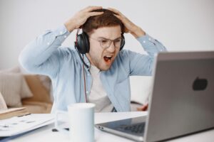 Hombre joven expresando frustración mientras trabaja con su ordenador portátil en una oficina en casa, representando la dificultad de conectar un teléfono al PC.