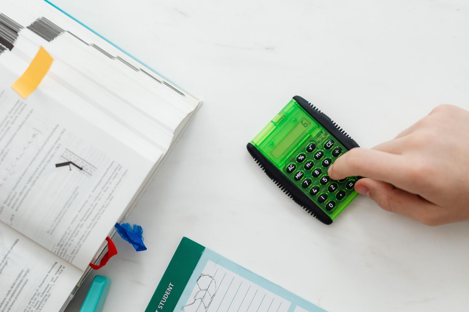 Primer plano de las manos de un alumno usando una calculadora verde junto a un libro de texto de matemáticas.